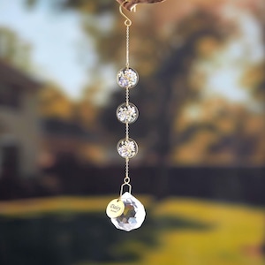 Puede incluir: Un atrapasoles colgante con tres esferas circulares transparentes de resina llenas de flores secas, conectadas por una cadena dorada. Una bola de cristal facetada y una etiqueta dorada con el nombre "Daisy" cuelgan debajo.