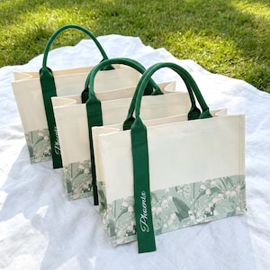 May include: Four off-white tote bags with green handles and accents. The bottom of each bag features a floral pattern with white flowers and green leaves. The word "Phoenix" is printed vertically on the green handle of each bag.