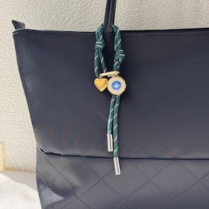 May include: A navy blue tote bag with a quilted base. A green and white rope charm with gold heart and eye pendant hangs from the handle. The charm has silver metal ends.