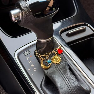 May include: A gold-toned chain bracelet featuring four charms: a crescent moon, a blue star, a sun with a face, and a red heart. The bracelet is wrapped around the black leather gear shift lever of an automatic vehicle with P R N D markings.