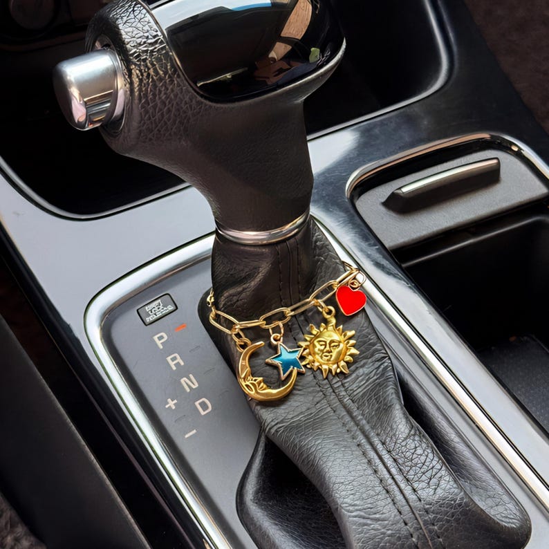 May include: A gold-toned chain bracelet featuring four charms: a crescent moon, a blue star, a sun with a face, and a red heart. The bracelet is wrapped around the black leather gear shift lever of an automatic vehicle with P R N D markings.