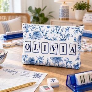 May include: A rectangular zipped pouch featuring a white background with a blue floral pattern and the name OLIVIA printed in dark blue letters, where each letter is stylised to resemble a mahjong tile on a white rectangular background.