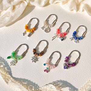 Peut inclure: Huit porte-clés en métal en forme de U ornés de perles colorées et de breloques argentées. Les breloques incluent une note de musique, une tasse à café, la lettre R, un signe du zodiaque, un cœur, la lettre N, un œil, une fleur, un soleil, des cerises et un éventail.