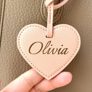 May include: A heart-shaped, light pink leather tag with the name "Olivia" engraved in a cursive font. The tag is attached to a light pink leather strap. The background is a textured, taupe-colored leather.