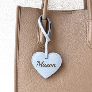 May include: A light blue heart-shaped tag with the name "Mason" in a cursive font is attached to a tan leather tote bag. The tag is suspended by a matching light blue loop. The bag has a textured surface and a top handle.