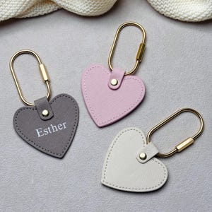 May include: Three heart-shaped keychains in gray, pink, and white. Each keychain has a gold-colored clasp and a small button. The gray keychain has the name "Esther" printed on it. The keychains are made of a textured material.