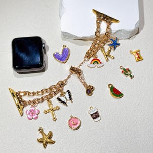 Op de afbeelding: Goudkleurige Apple Watch-band met diverse bedels, waaronder een kruis, een vlinder, een bloem en een regenboog. Andere bedels zijn een koffiekopje, een watermeloen en een paars hart. De band wordt gepresenteerd op een wit oppervlak.