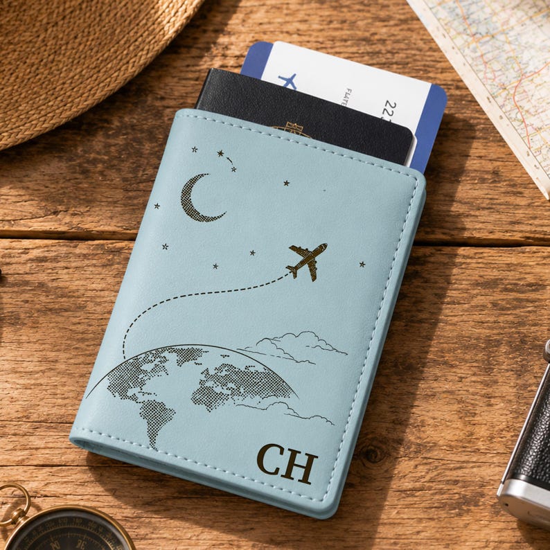 May include: A light blue passport holder featuring a black graphic print of a crescent moon, stars, a globe, and an airplane with a dotted flight path. The initials CH are printed in black at the bottom, and a black passport is inserted inside.