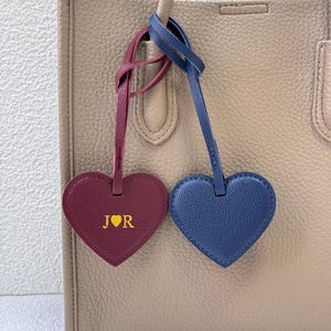 Può includere: Una borsa tote beige con due ciondoli a forma di cuore in pelle. Un ciondolo è bordeaux con la scritta dorata "J ♥ R", l'altro è blu. I ciondoli sono attaccati al manico della borsa.