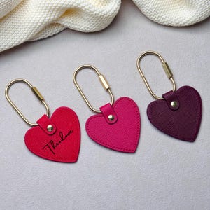 May include: Three heart-shaped keychains with gold-toned oval rings. The left keychain is bright red with the name Theodore printed in black script. The middle keychain is fuchsia, and the right keychain is deep purple. Each features a gold rivet.