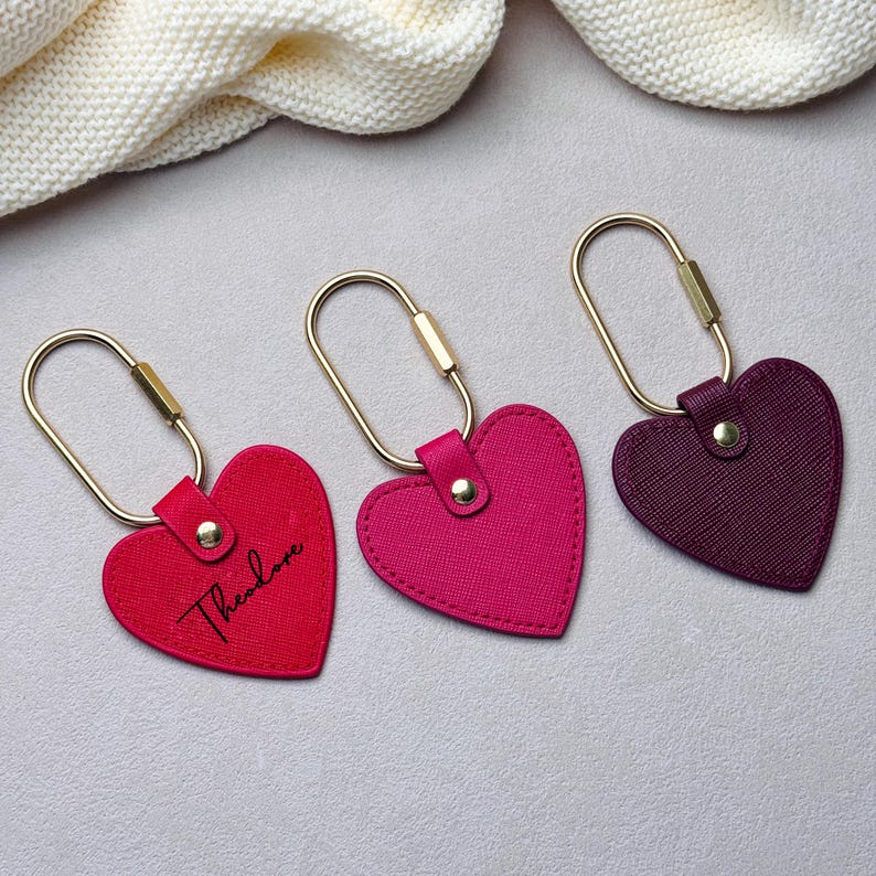 May include: Three heart-shaped keychains with gold-toned oval rings. The left keychain is bright red with the name Theodore printed in black script. The middle keychain is fuchsia, and the right keychain is deep purple. Each features a gold rivet.