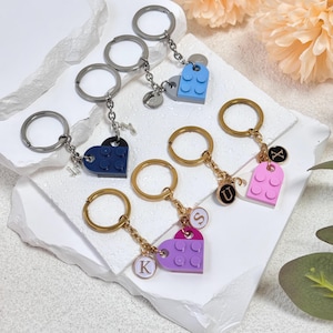 May include: Keychains with heart-shaped charms in blue, navy, purple, and pink. Each keychain has a metal ring and chain, with a small charm featuring a letter. The charms are designed to resemble interlocking toy bricks.