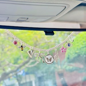 Puede incluir: Amuleto de coche plateado con varios dijes colgantes, que incluyen una gema rosa, una abeja, un corazón, un lazo, una mariposa, una clave de sol, una flor rosa, un ojo turco y una estrella. Cuelga del espejo retrovisor.
