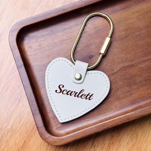 May include: A heart-shaped keychain with the name "Scarlett" in a cursive font. The keychain is made of light beige leather and has a gold-colored metal clasp. The keychain is resting on a wooden tray.