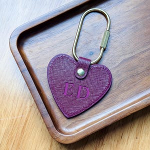 May include: A heart-shaped, plum-colored leather keychain with the initials "ED" embossed on the front. It features a gold-colored metal clasp and a small leather strap. The keychain is resting on a wooden tray.
