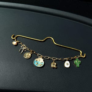 以下が含まれることがあります： ゴールドカラーの安全ピンとチャームのチェーン。チャームには、リボン、貝殻、「R」の文字、太陽、緑色のサボテンが含まれます。貝殻のチャームは青とターコイズのエナメルです。ピンは黒い表面にあります。