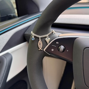 May include: A silver charm bracelet featuring a blue crystal, a heart, and a fish pendant inscribed with the word "JESUS". The bracelet is shown on a black steering wheel.