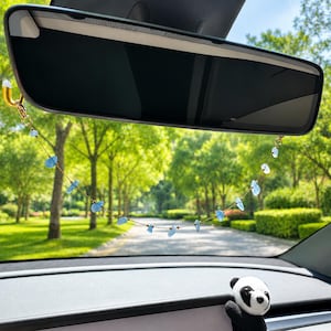 May include: A car rearview mirror charm featuring a delicate gold-toned chain adorned with small, irregular, light blue translucent stone beads. The chain is attached to the mirror frame, hanging in a gentle curve against the view of a green park.