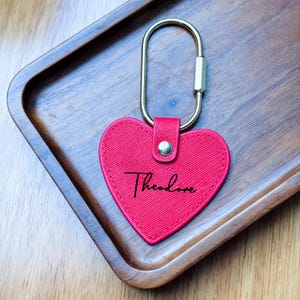 May include: A red heart-shaped keychain with the name "Theodore" in cursive script. The keychain has a gold-colored clasp and a small, gold-colored rivet. The keychain is on a wooden tray.