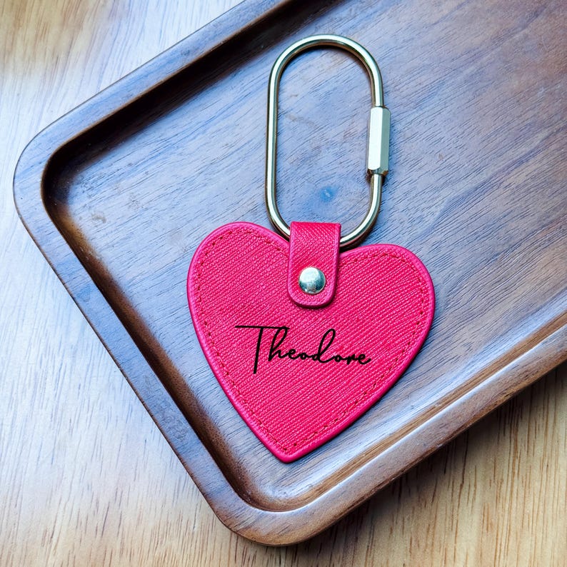May include: A red heart-shaped keychain with the name "Theodore" in cursive script. The keychain has a gold-colored clasp and a small, gold-colored rivet. The keychain is on a wooden tray.
