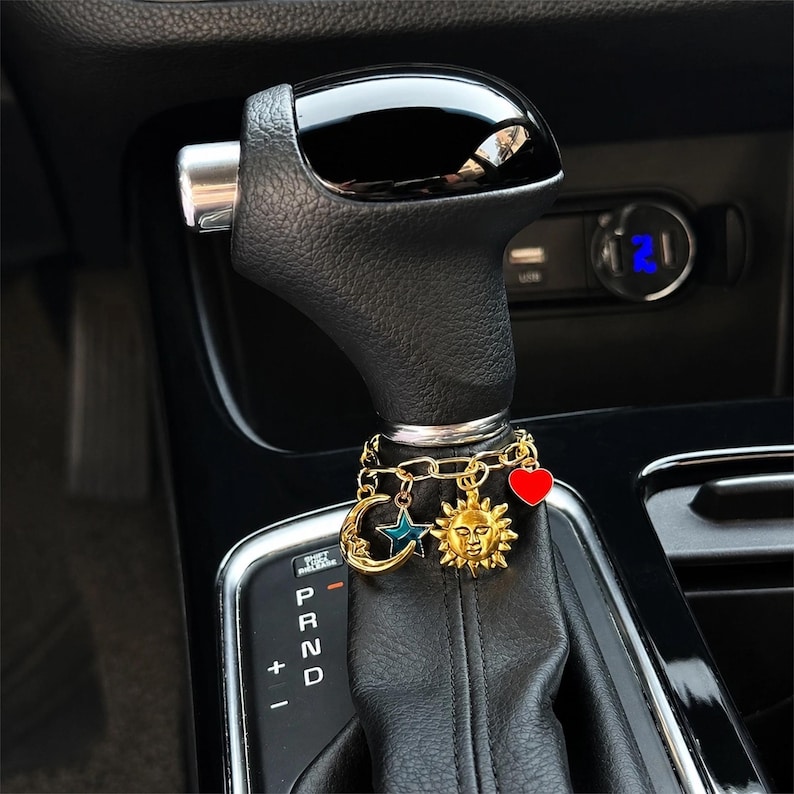 A stylish custom gear shift chain charm wrapped around a car shifter, featuring gold-tone chains and assorted charms including a moon, star, sun face, and red heart. A cute personalized car interior accessory for adding sparkle, color, and personality to your vehicle.