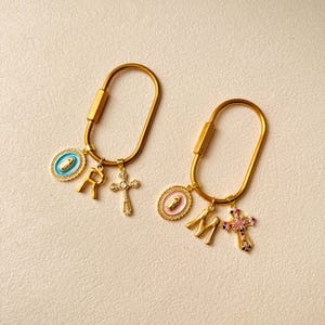 May include: Two gold-colored keychains with religious charms. Each keychain has a carabiner-style clasp and features a cross pendant, a letter charm, and a medallion with a religious image. One keychain has the letter "R", the other has the letter "M".