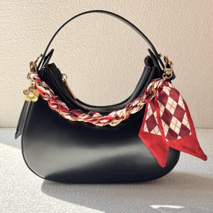 May include: Black handbag with a curved design, adorned with a gold chain and a red and white patterned scarf. The bag has a single handle and a gold flower charm. The bag is made of a smooth material.