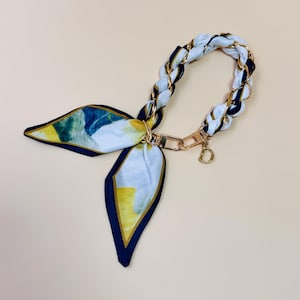 Puede incluir: Un pa&ntilde;uelo estampado en blanco, azul y amarillo, con un asa de cadena dorada. Cuenta con un cierre dorado y un peque&ntilde;o dije dorado con la letra "D". El fondo es beige.