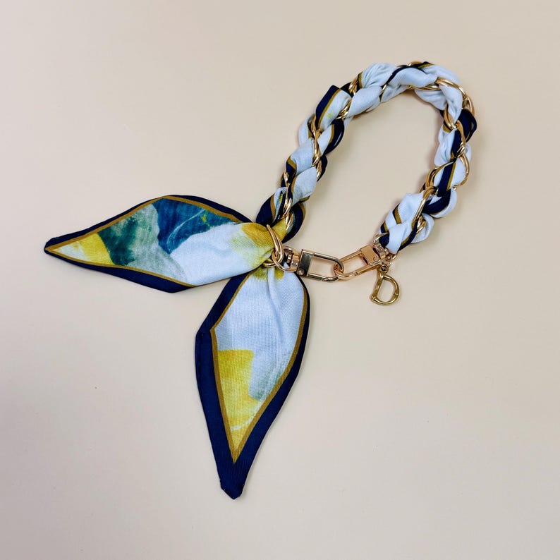 Puede incluir: Un pa&ntilde;uelo estampado en blanco, azul y amarillo, con un asa de cadena dorada. Cuenta con un cierre dorado y un peque&ntilde;o dije dorado con la letra "D". El fondo es beige.