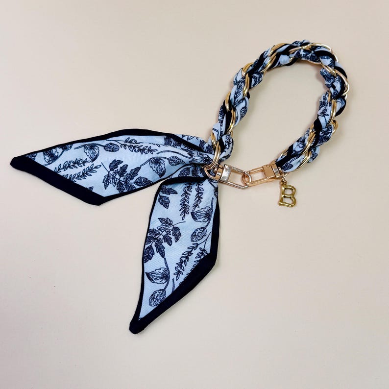 Puede incluir: Un pa&ntilde;uelo con estampado azul claro y negro con un asa de cadena dorada. El pa&ntilde;uelo presenta un dise&ntilde;o floral y un borde negro. Un dije dorado con la letra "B" cuelga de la cadena.