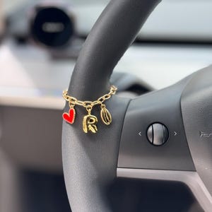 Op de afbeelding: Gouden bedelarmband met een rood hart, de letter "R" en een schelp. De armband is bevestigd aan een zwart stuurwiel. De bedels zijn gedetailleerd met rood email.