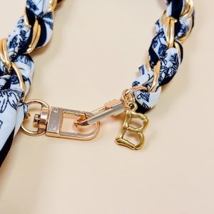 Puede incluir: Una correa de cadena dorada con una bufanda con estampado floral en blanco y negro tejida a trav&eacute;s. La correa presenta un cierre dorado y un dije con la letra "B" dorada, ideal para un bolso o accesorio.