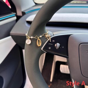 May include: A gold-colored charm bracelet with a variety of charms, including a shell, a flower, a heart, and a disc with the letter "F". The bracelet is attached to a car's steering wheel.