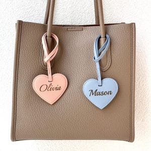 Two heart-shaped leather bag charms in pink and blue, laser engraved with the names “Olivia” and “Mason,” attached to a taupe handbag.