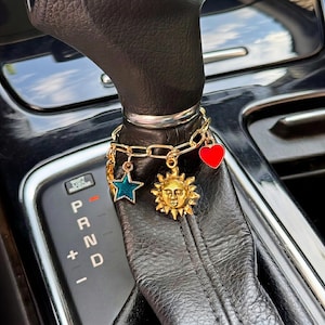 May include: A gold-toned chain bracelet wrapped around a black leather car gear shift knob. The bracelet features three dangling charms: a teal enamel star, a gold sun face with rays, and a solid red heart charm with a smooth, glossy finish.