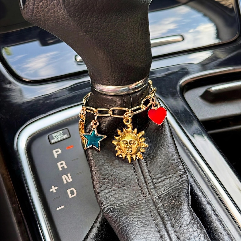May include: A gold-toned chain bracelet wrapped around a black leather car gear shift knob. The bracelet features three dangling charms: a teal enamel star, a gold sun face with rays, and a solid red heart charm with a smooth, glossy finish.