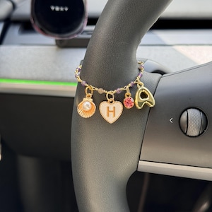 Peut inclure: Bracelet de charme doré avec des breloques en forme de coquillage, de cœur avec la lettre "H", de gemme rose et de lettre "A". Le bracelet, orné de perles violettes, est suspendu à un volant de voiture.