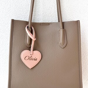 May include: A taupe-colored tote bag with long handles and a pink heart-shaped tag. The tag is inscribed with the name "Olivia" in a cursive font. The bag has a rectangular shape and a textured surface.