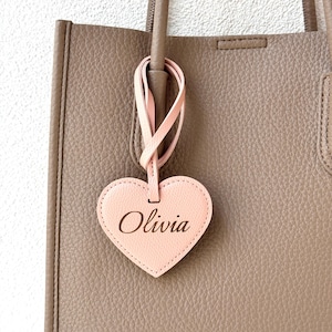 May include: A light pink heart-shaped tag with the name "Olivia" in a cursive font, hanging from a taupe tote bag. The tag is attached to the bag by a pink leather strap. The bag has a textured surface.