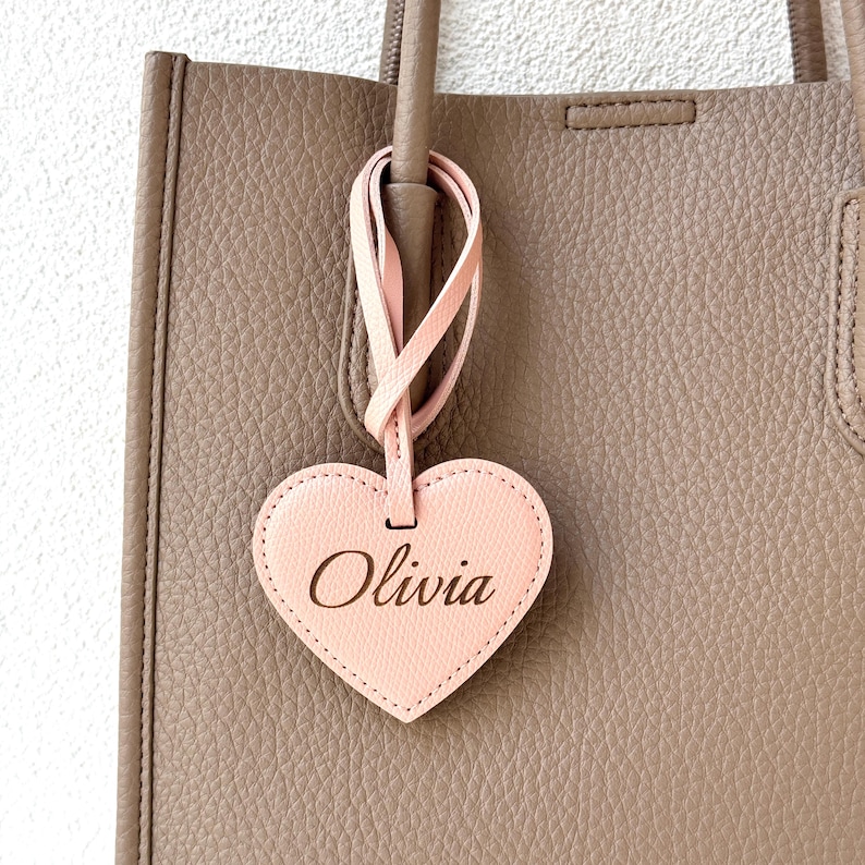May include: A light pink heart-shaped tag with the name "Olivia" in a cursive font, hanging from a taupe tote bag. The tag is attached to the bag by a pink leather strap. The bag has a textured surface.