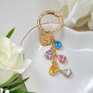 May include: Gold-toned keychain with a circular clasp and a small square charm engraved with the letter "S". The keychain features a chain with five teardrop-shaped, colorful gemstones: blue, purple, pink, yellow, and white.