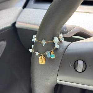May include: A gold-toned chain bracelet featuring light blue, irregular-shaped stone beads, a rectangular gold charm engraved with the letter R, and a small circular charm with a textured, bright turquoise center, arranged in a double-strand style.