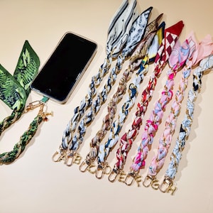 Puede incluir: Un tel&eacute;fono inteligente con una funda transparente est&aacute; conectado a un cord&oacute;n trenzado verde y dorado con una bufanda con estampado bot&aacute;nico. Se muestran varios otros cordones trenzados con bufandas estampadas y dijes de iniciales doradas.