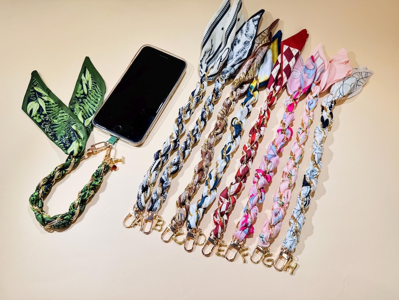 Puede incluir: Un tel&eacute;fono inteligente con una funda transparente est&aacute; conectado a un cord&oacute;n trenzado verde y dorado con una bufanda con estampado bot&aacute;nico. Se muestran varios otros cordones trenzados con bufandas estampadas y dijes de iniciales doradas.