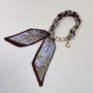 Puede incluir: Un pa&ntilde;uelo decorativo con un asa de cadena trenzada. El pa&ntilde;uelo presenta un estampado floral en tonos morado, marr&oacute;n y dorado. Un dije dorado con la letra "C" cuelga de la cadena.