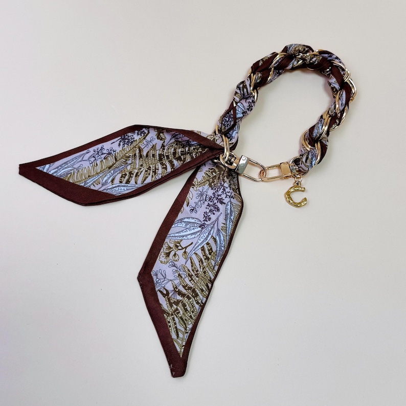 Puede incluir: Un pa&ntilde;uelo decorativo con un asa de cadena trenzada. El pa&ntilde;uelo presenta un estampado floral en tonos morado, marr&oacute;n y dorado. Un dije dorado con la letra "C" cuelga de la cadena.