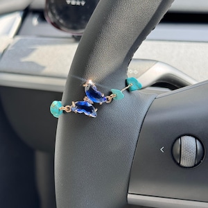 Op de afbeelding: Een armband met een centrale vlinderbedel van gefacetteerd blauw glas, verbonden door goudkleurige metalen schakels met verschillende onregelmatige, lichtblauwe turkooiskleurige stenen kralen. Het ontwerp is delicaat en symmetrisch.