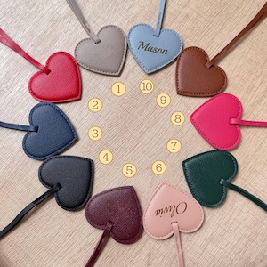 May include: A collection of heart-shaped leather tags in various colors, including red, blue, and pink, arranged in a circular pattern. One tag is light blue with the name "Mason" and another is pink with the name "Olivia". Each tag has a matching strap.