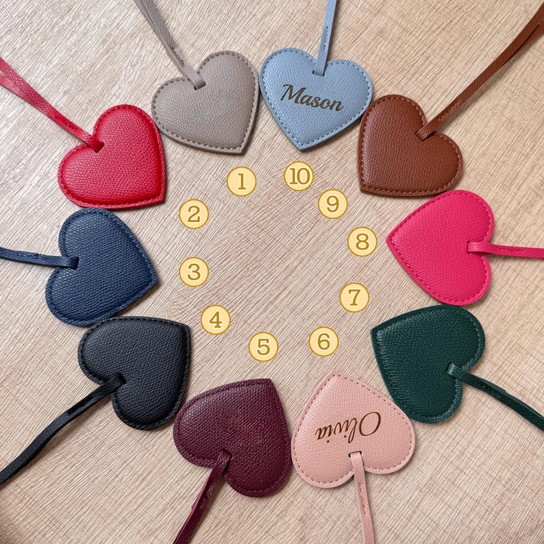 May include: A collection of heart-shaped leather tags in various colors, including red, blue, and pink, arranged in a circular pattern. One tag is light blue with the name "Mason" and another is pink with the name "Olivia". Each tag has a matching strap.