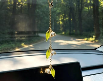 Könnte beinhalten: Ein goldfarbener Autoanhänger mit drei Maiglöckchen-Blütenanhängern mit weißen Blüten und grünen Blättern. Der Anhänger hängt an einer Kette vor einem verschwommenen Waldbild.